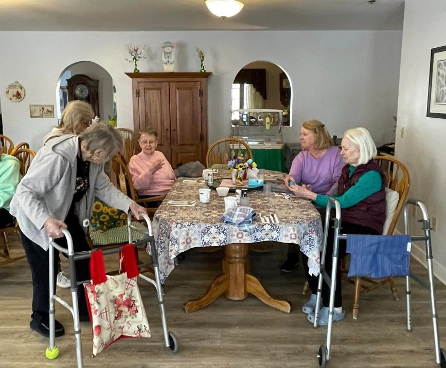 A group of elderly women gathered around a table, engaging in conversation at a nursing home.