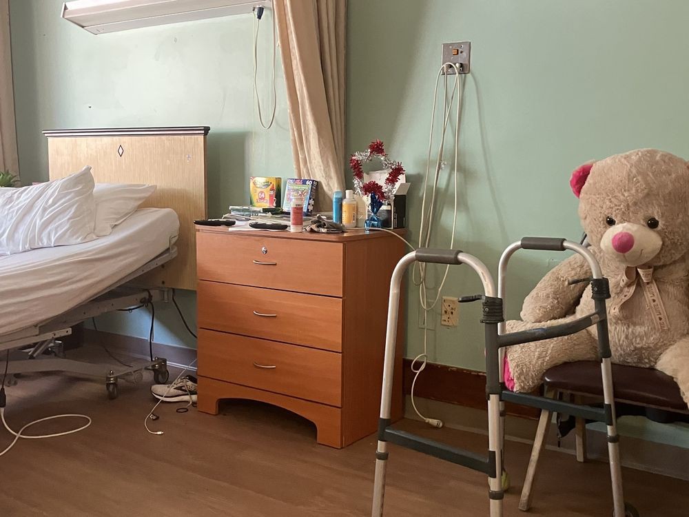 A hospital room with a bed, a dresser with personal items, and a walker with a teddy bear resting on it.