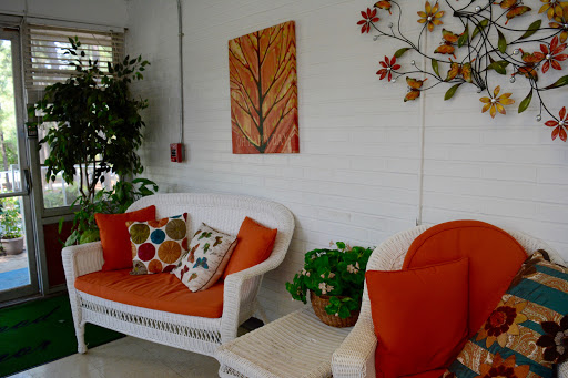 A cozy living room featuring a white wicker sofa with orange cushions, colorful patterned pillows, and decorative wall art.