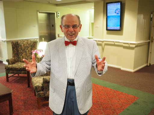 A cheerful man in a bow tie and light suit, expressing joy in an indoor setting.