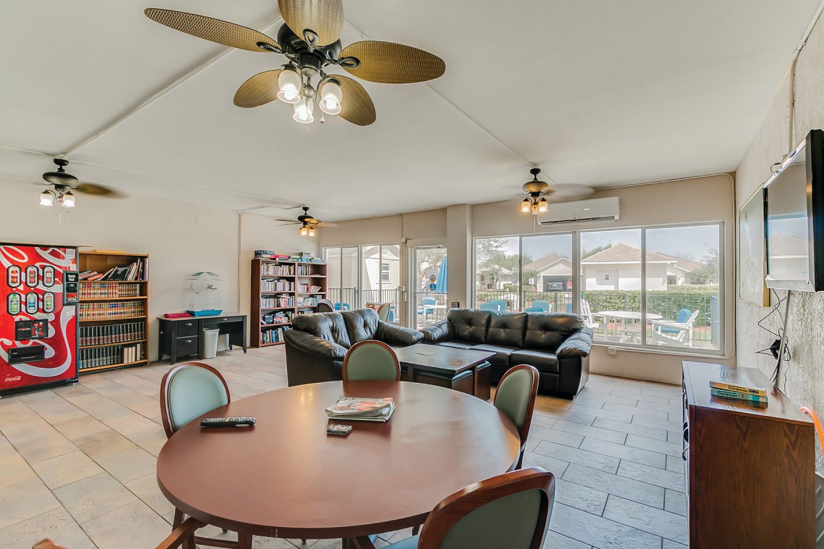 A spacious living room with large windows, featuring a dining table, comfortable sofa, ceiling fans, and a bookshelf.