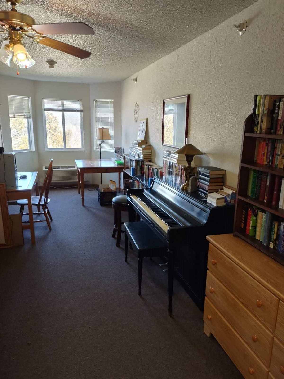 A cozy living room with a piano, bookshelf, and workspace by the window.