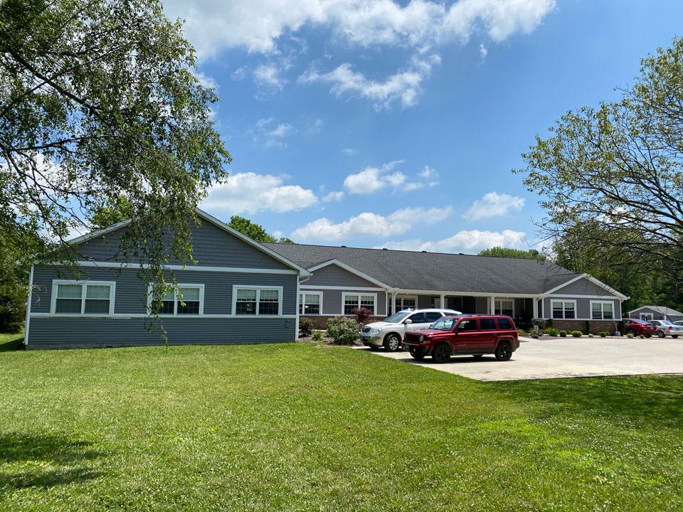 A suburban house with a well-maintained lawn, two cars parked in the driveway under a blue sky with scattered clouds.