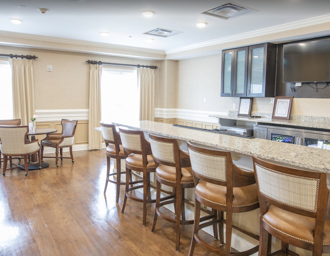 A spacious kitchen area featuring a long countertop with bar stools and wooden flooring, complemented by cabinetry and natural lighting.