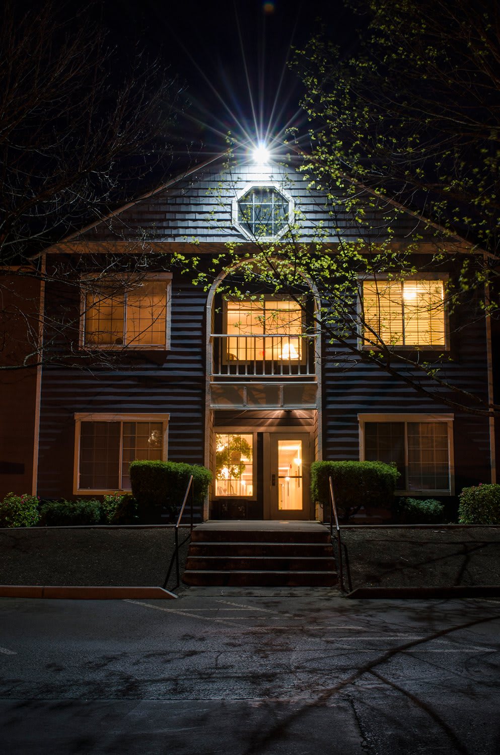A house at night with illuminated windows and entrance, surrounded by trees.