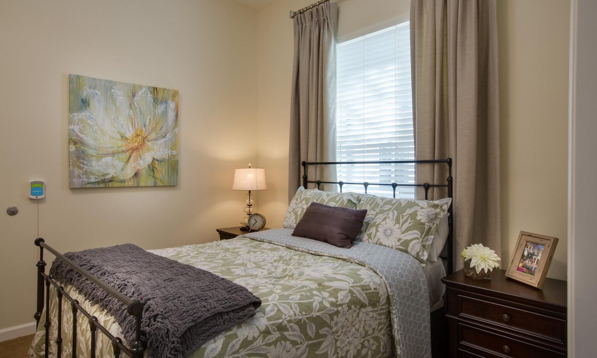 A cozy bedroom with a neatly made bed, a lamp on the nightstand, and decorative curtains by the window.