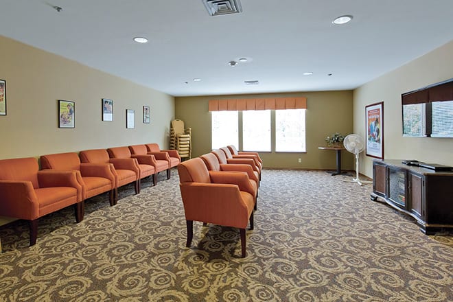 An empty waiting room featuring rows of orange chairs and a patterned carpet.