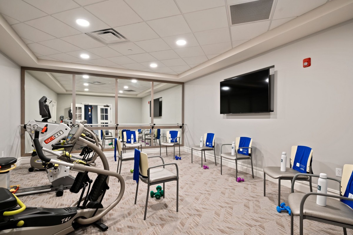 A well-lit exercise room with chairs arranged in a circle, featuring a large television and mirrored walls.