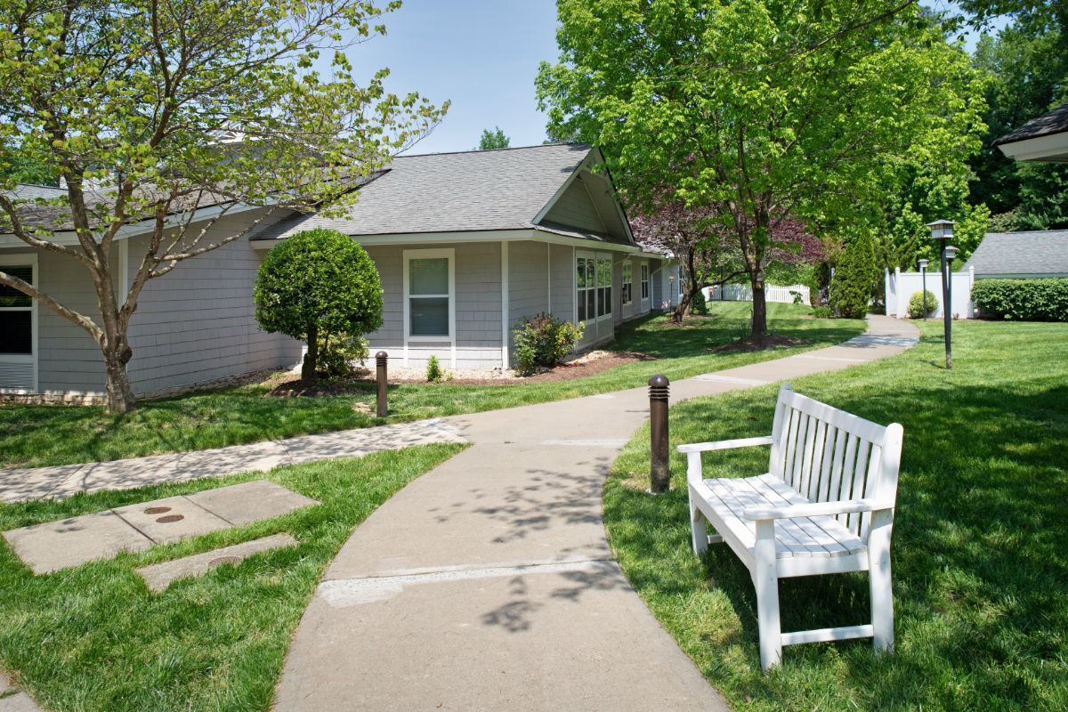 A peaceful suburban scene with a white bench along a pathway, surrounded by trees and a small house.