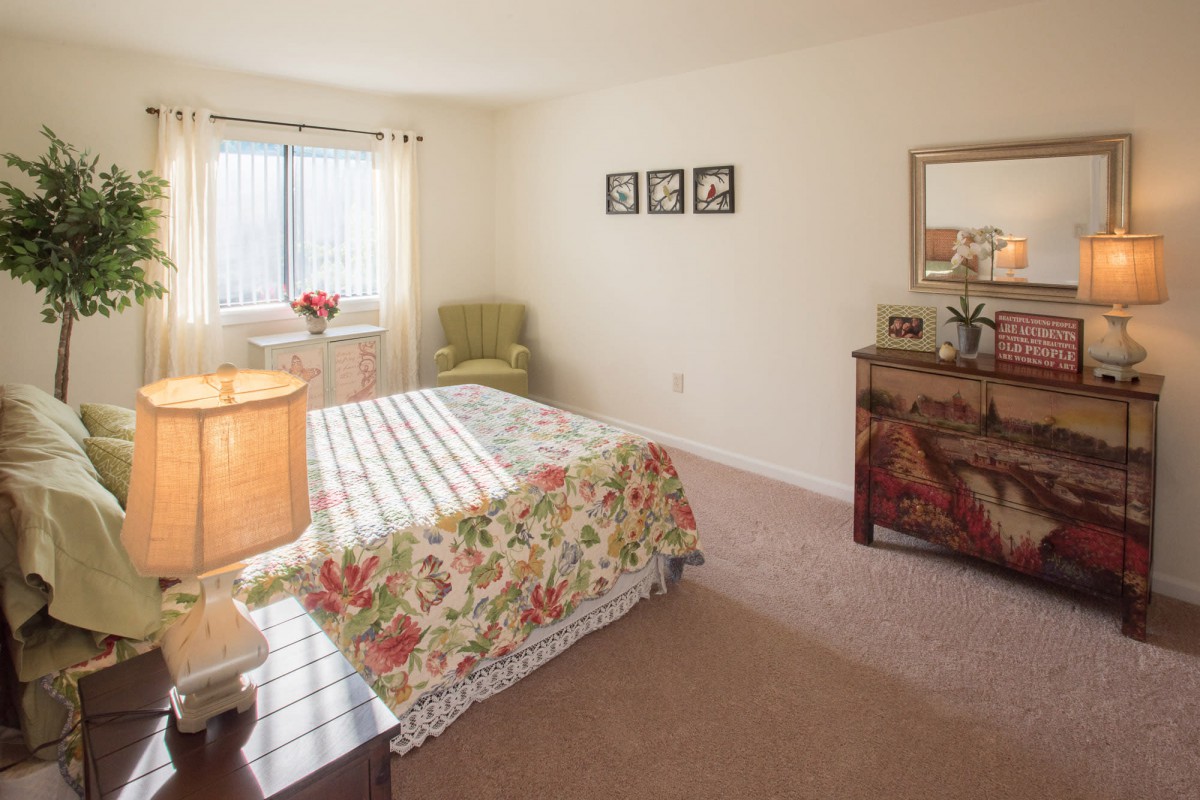 A cozy bedroom with floral bedspread, dresser, and window letting in natural light.