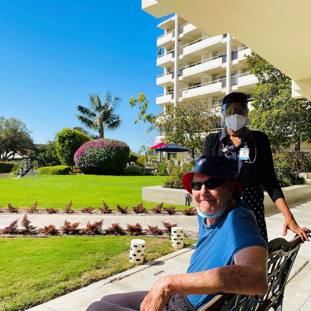 A man in a wheelchair enjoys a sunny day outdoors with a caregiver in a garden setting.