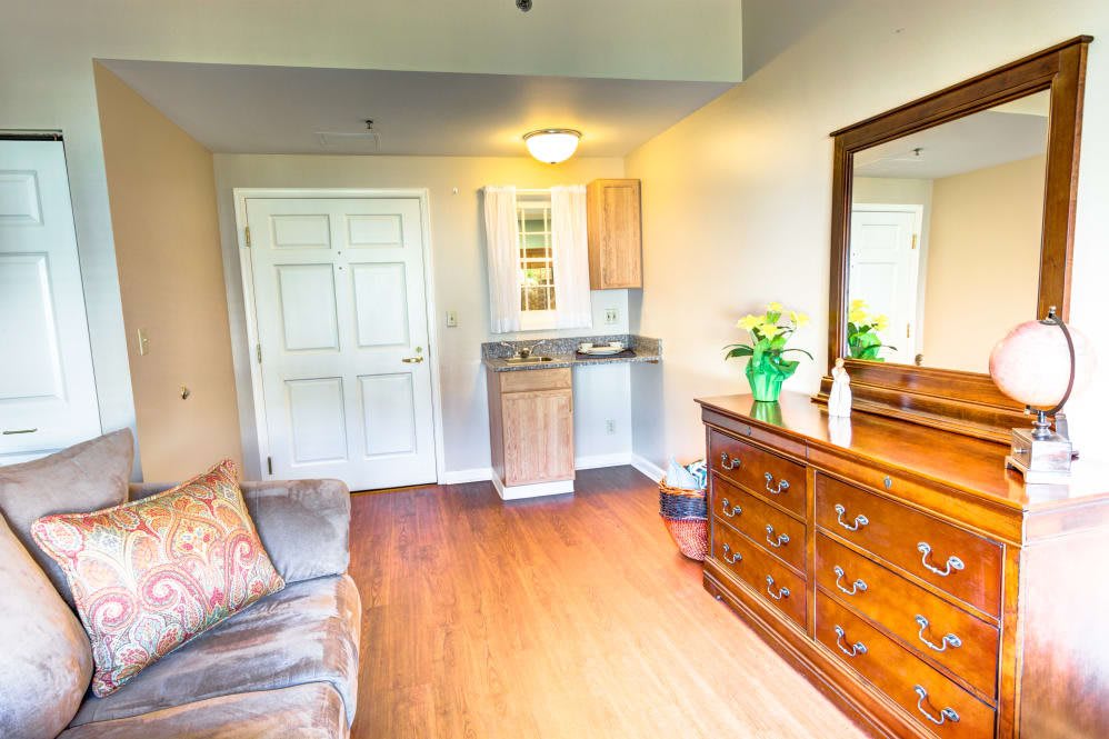 A cozy living room with a sofa, dresser, mirror, and vibrant flowers on a wooden floor.