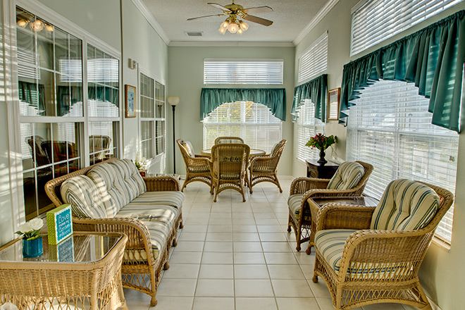 A bright sunroom featuring wicker furniture and a dining area, with large windows and a ceiling fan.