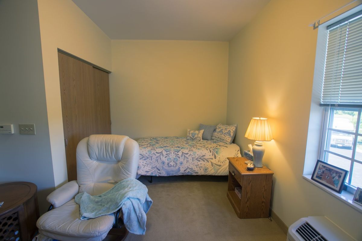 A cozy bedroom with a bed, chair, and nightstand featuring a lamp.