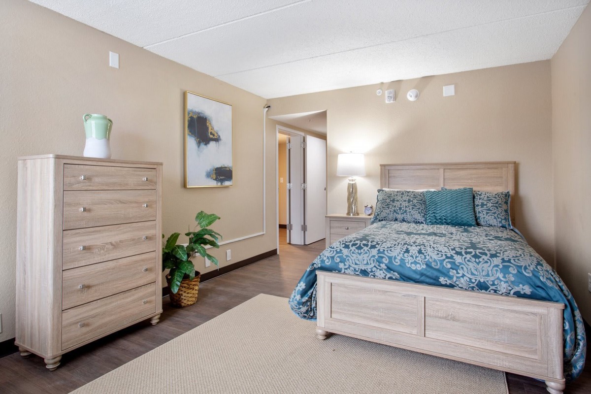 A cozy bedroom with stylish decor, featuring a bed with blue bedding, a wooden dresser, potted plant, and wall art.