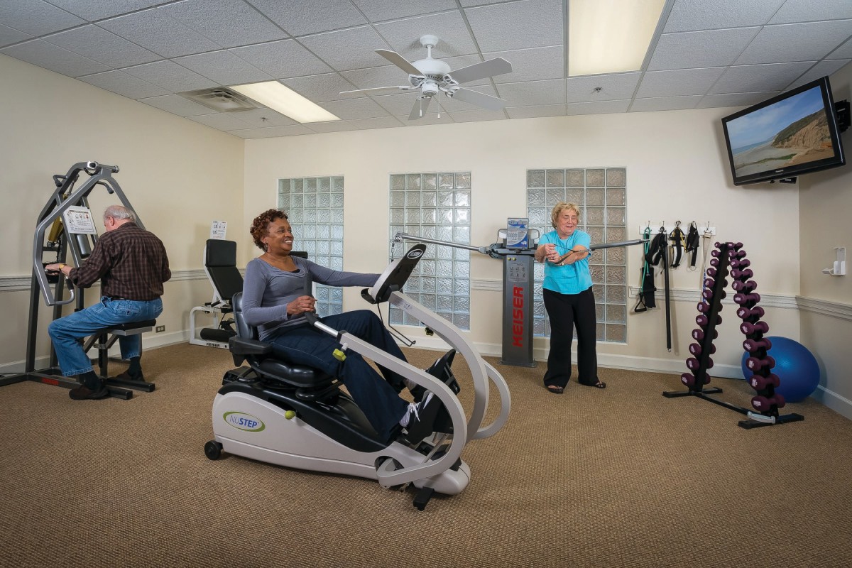 A group of people exercising indoors with fitness equipment, including a recumbent bike.