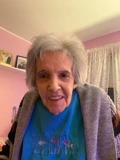 An elderly woman with grey hair smiles warmly inside a cozy room.