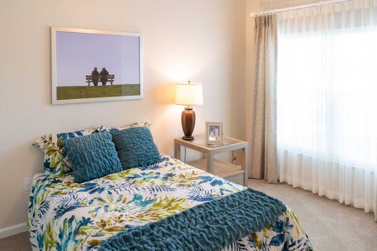 A cozy bedroom with a floral bedspread, teal pillows, and a bedside lamp on a nightstand.