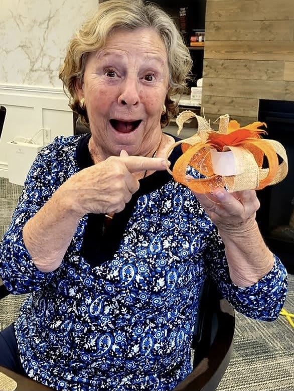 An elderly woman joyfully displays her handmade pumpkin craft indoors.