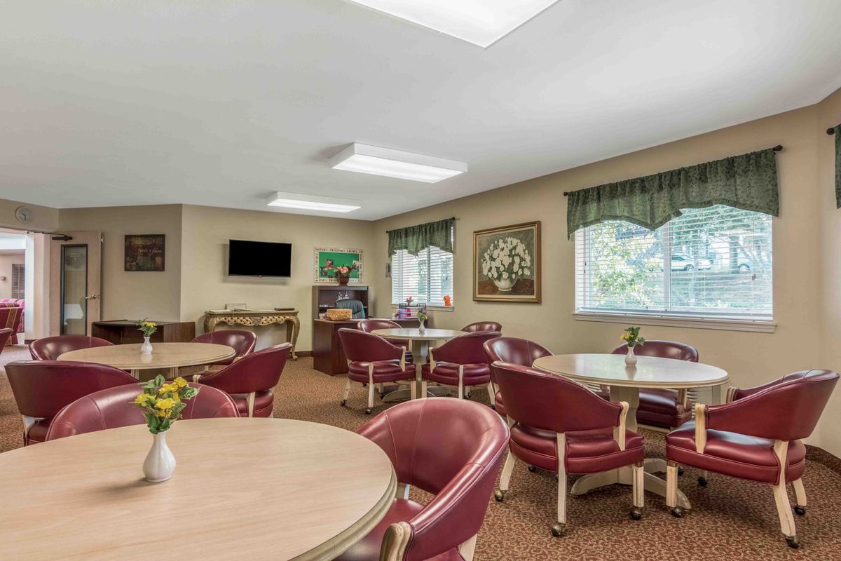 A well-lit dining area with round tables and red chairs, featuring a television and artwork on the walls.