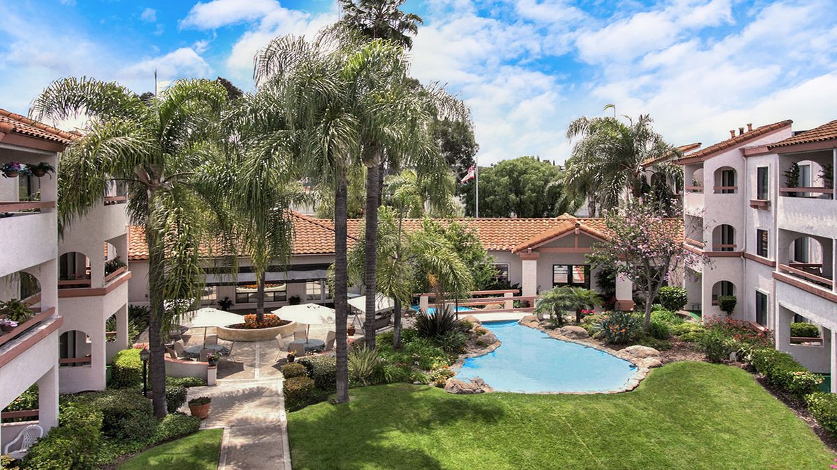 A serene outdoor setting featuring a swimming pool surrounded by lush greenery and palm trees, adjacent to a house with a terracotta roof.