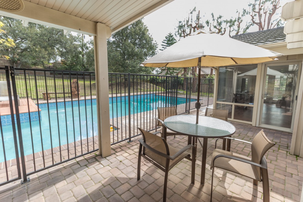 A cozy patio area with a table and chairs overlooks a backyard pool, surrounded by a safety fence and shaded by an umbrella.