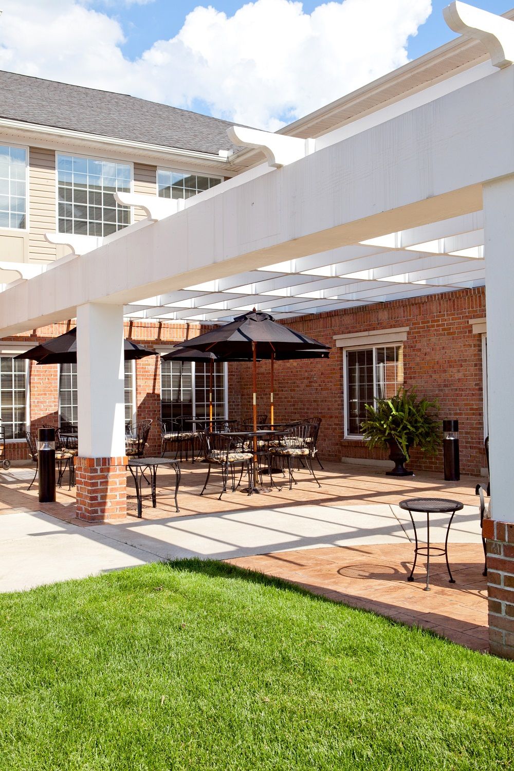 A sunny patio area with outdoor seating and umbrellas under a pergola, adjacent to a brick building.