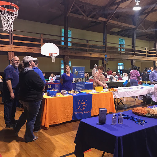 A lively community event taking place in a gymnasium, featuring various vendor tables and attendees interacting.