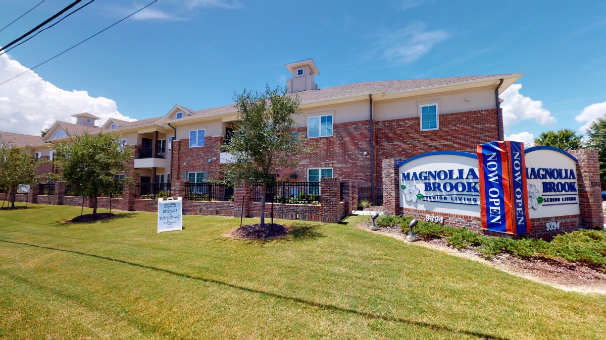 A brick apartment complex with a sign in front reading 'Magnolia Blooms' and well-maintained landscaping.