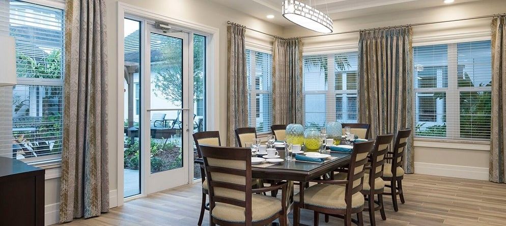 A modern dining room with a large table and chairs, surrounded by windows allowing in natural light.