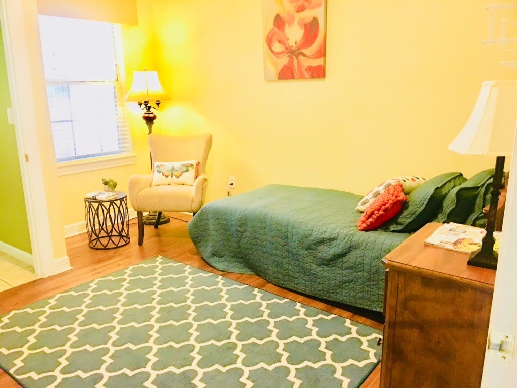 A cozy bedroom with a green bedspread, patterned rug, and a soft armchair near a lamp.