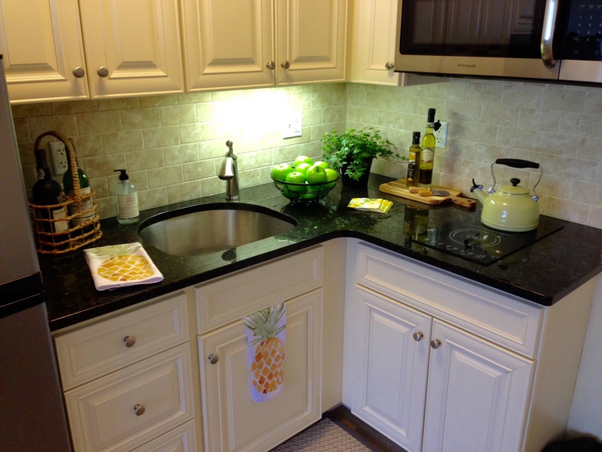 A cozy kitchen corner with cream cabinets and a black countertop, featuring a sink, herbs, and essential appliances.