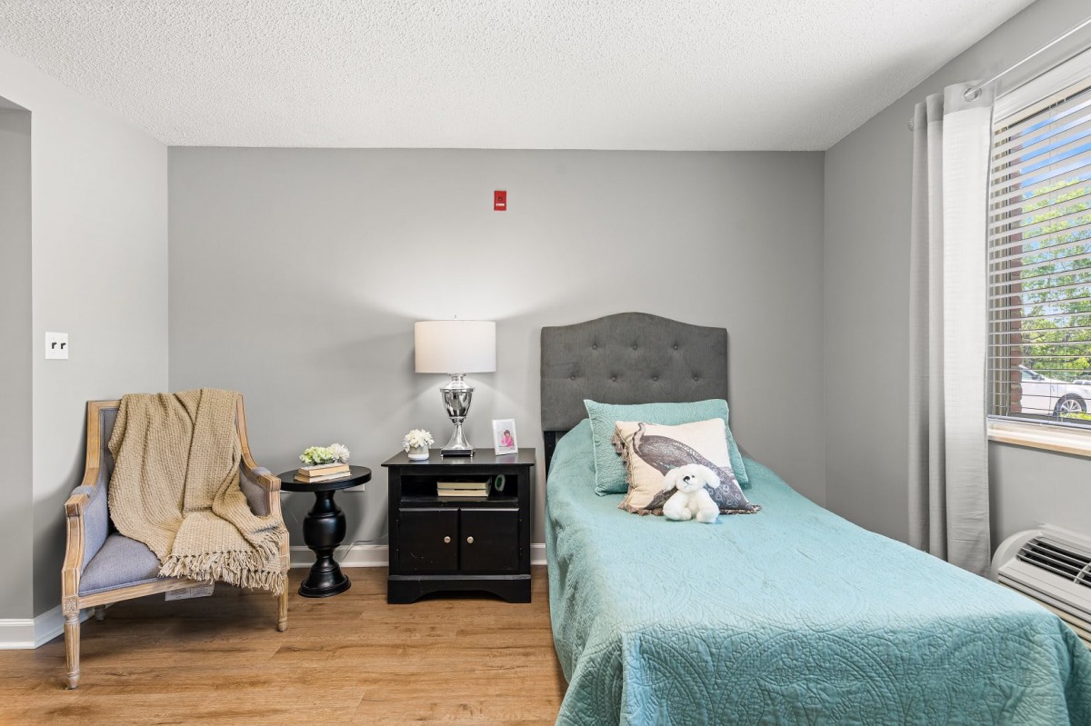 A cozy bedroom with a single bed and nightstand, featuring a lamp and decorative pillows.