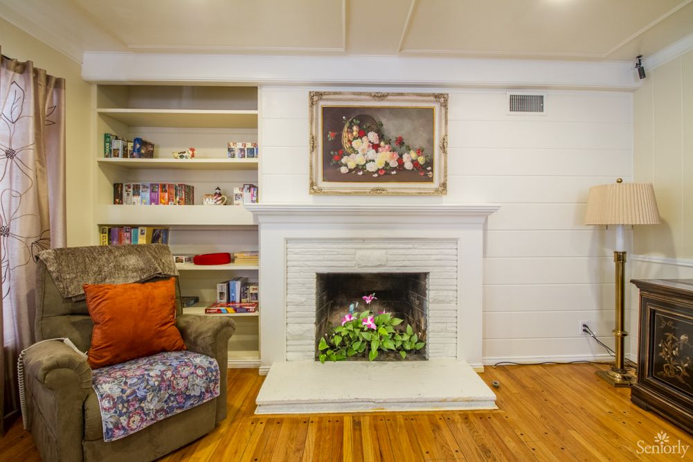 A cozy living room featuring a white fireplace with surrounding bookshelves, adorned with artwork and complemented by a comfy armchair and wooden flooring.
