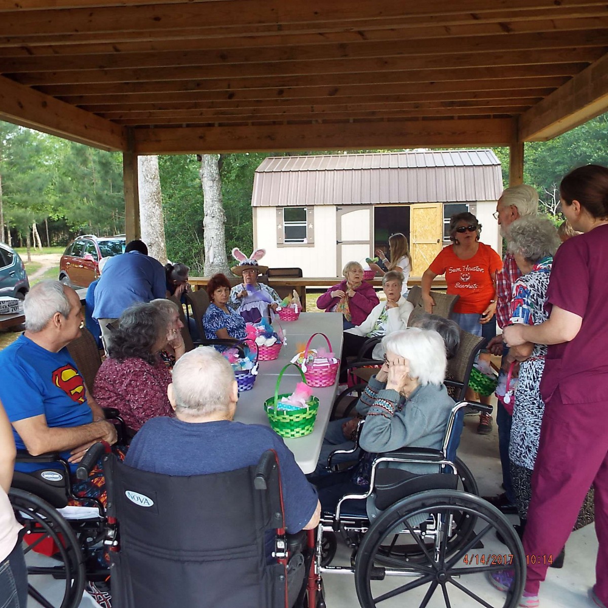 A group of people, including seniors in wheelchairs, enjoy an outdoor picnic under a wooden shelter.