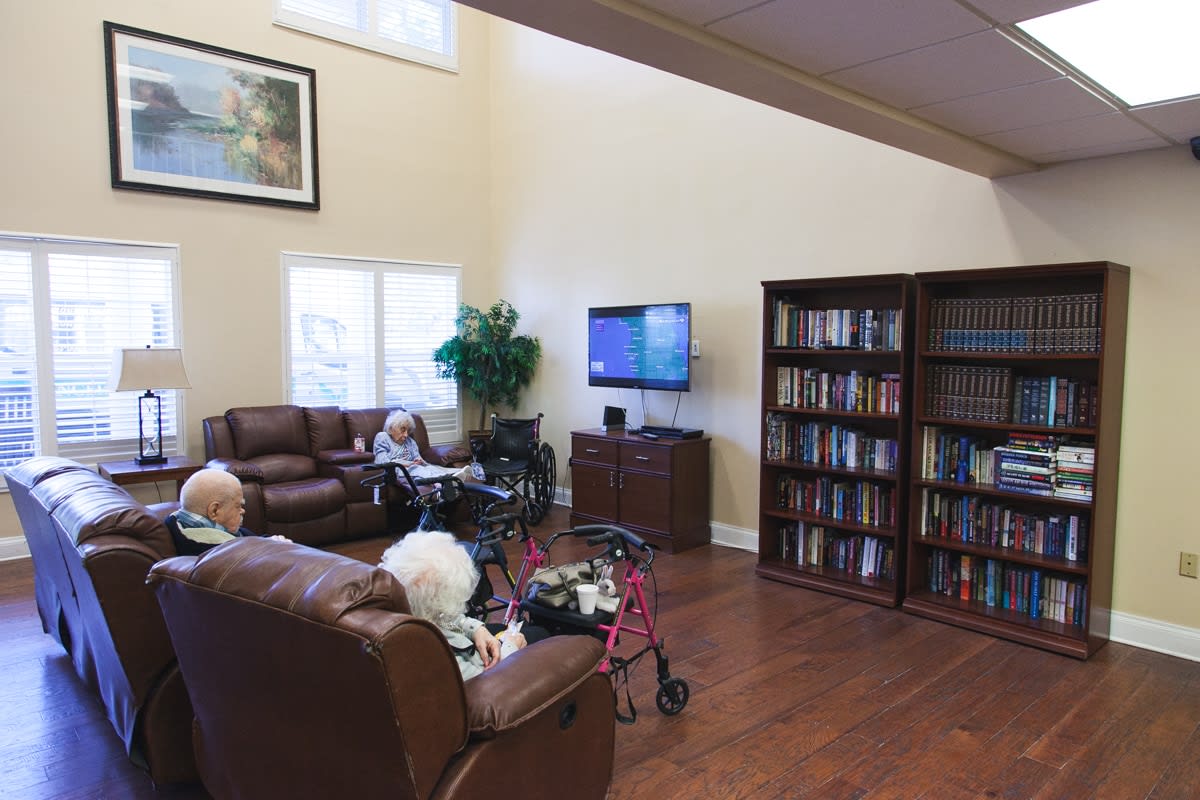 A cozy living room with seniors seated on leather sofas, surrounded by bookshelves and a television.