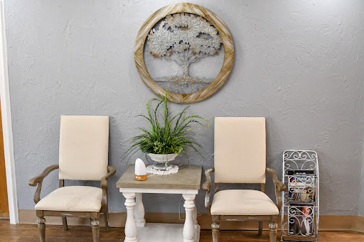 A cozy seating area featuring two beige chairs and a decorative table, complemented by a wall-mounted tree art piece and a potted plant for a touch of nature.