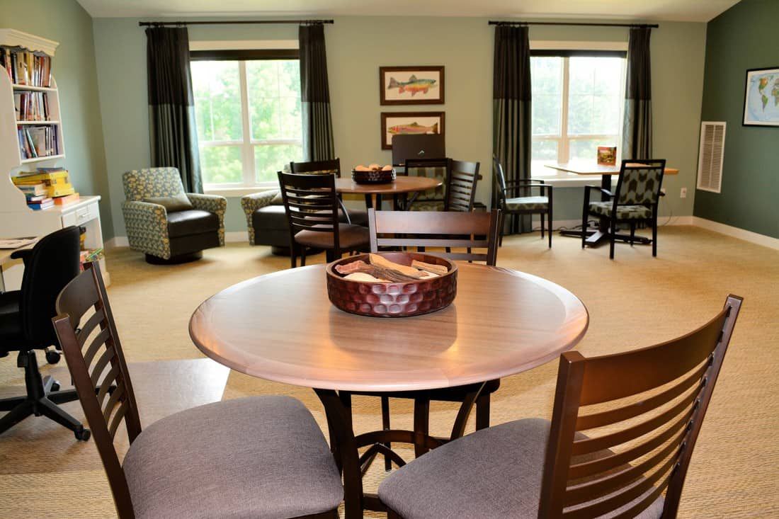 A cozy living room with multiple seating areas, featuring tables and chairs arranged near large windows.