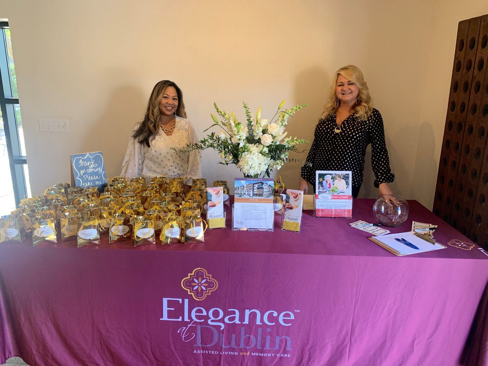 Two women at a business event stand behind a table with a purple Elegance in Design tablecloth, featuring cookies, brochures, and a floral arrangement.