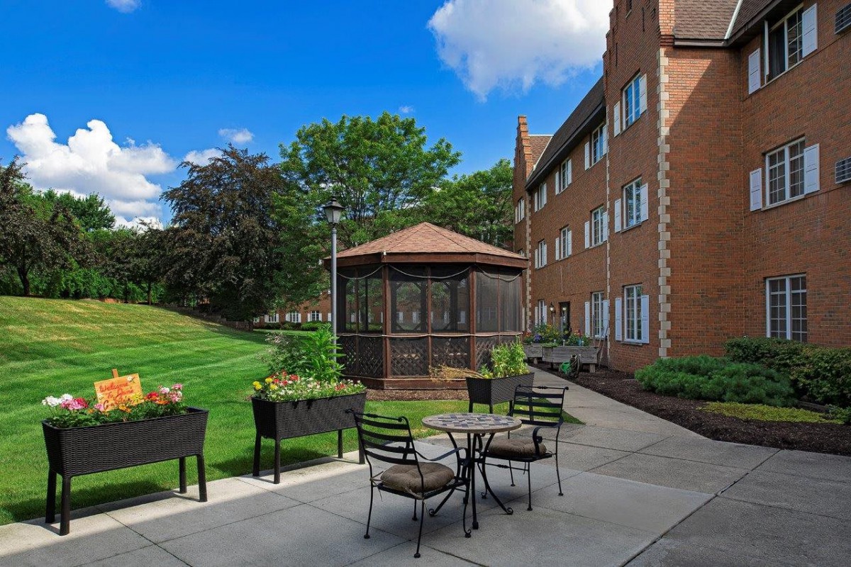 A peaceful outdoor patio area next to an apartment building with seating, a gazebo, and surrounding greenery.