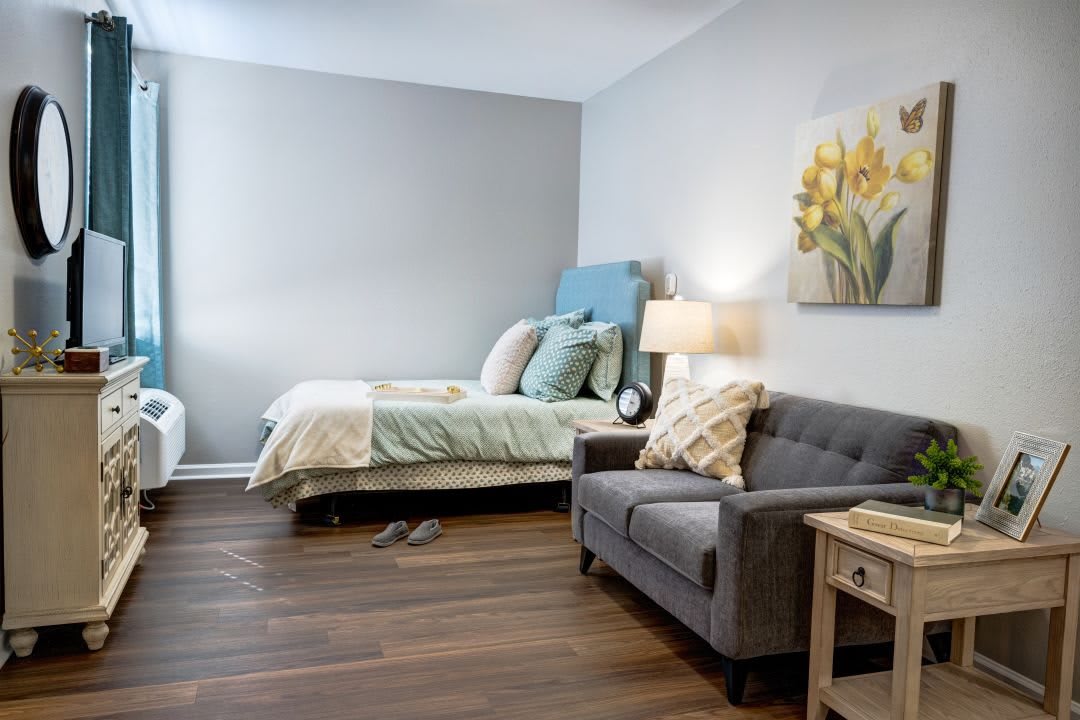 A cozy bedroom featuring a neatly made bed with a blue headboard, a grey sofa, and a side table with a lamp. The room has hardwood floors and is decorated with floral artwork on the wall.
