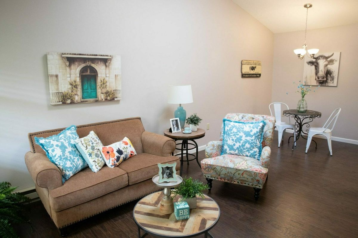 A cozy living room with a brown sofa, patterned armchair, and decorative pillows. A coffee table is centered, adorned with decor items, and framed artwork hangs on the walls.
