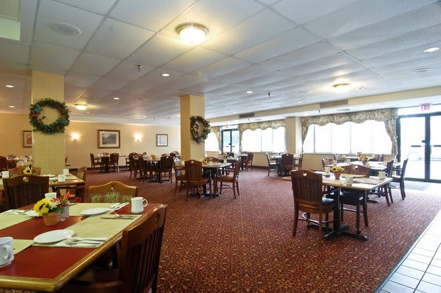 A spacious restaurant dining area with neatly arranged tables and chairs, featuring warm lighting and a carpeted floor.
