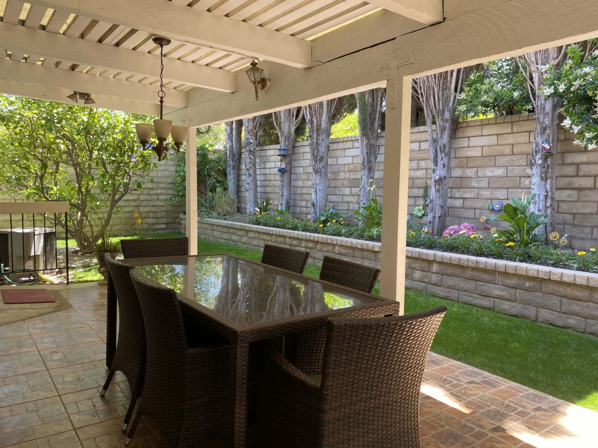 A cozy patio setup with a dining table and chairs under a pergola, surrounded by a lush garden.