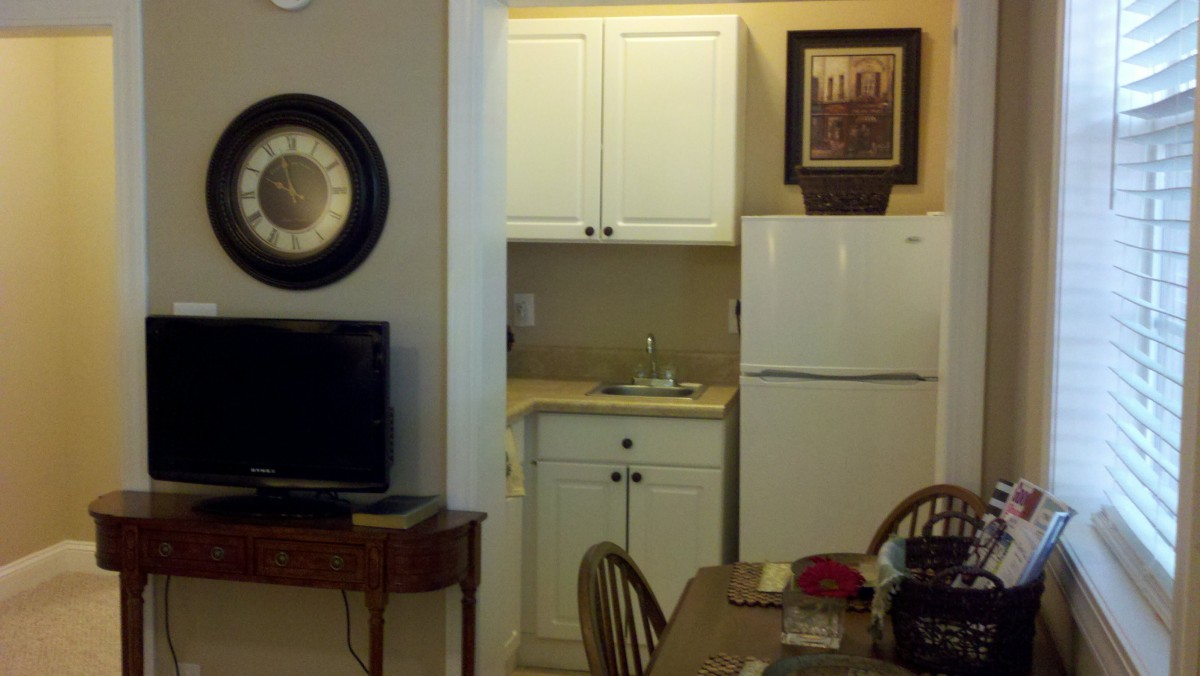 A small kitchen area with white cabinets, a refrigerator, and a nearby dining table. A TV and artwork are also visible.