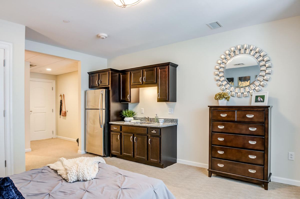 A cozy bedroom with a kitchenette, featuring brown cabinets and a stainless steel fridge.
