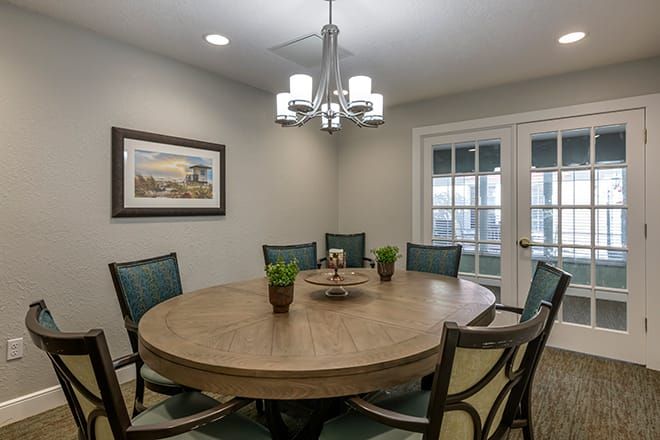 A cozy dining room with a round wooden table and upholstered chairs, illuminated by a modern chandelier. Small plants and artwork add to the decor beside elegant French doors.