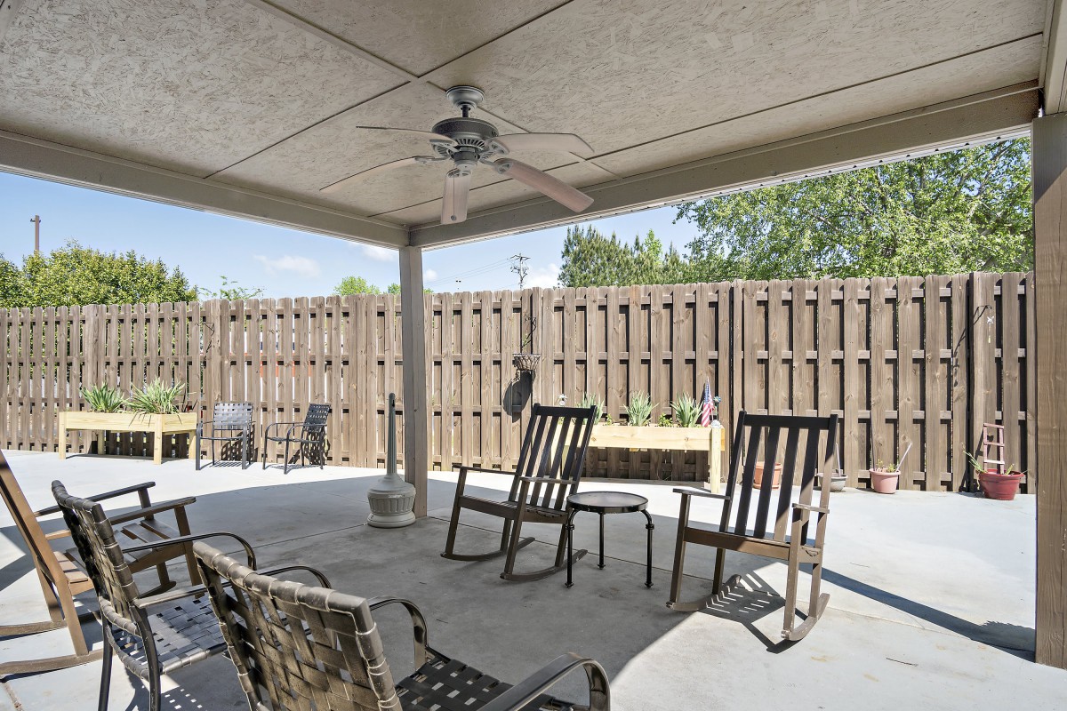 A covered patio area with seating and a ceiling fan, surrounded by a wooden fence.