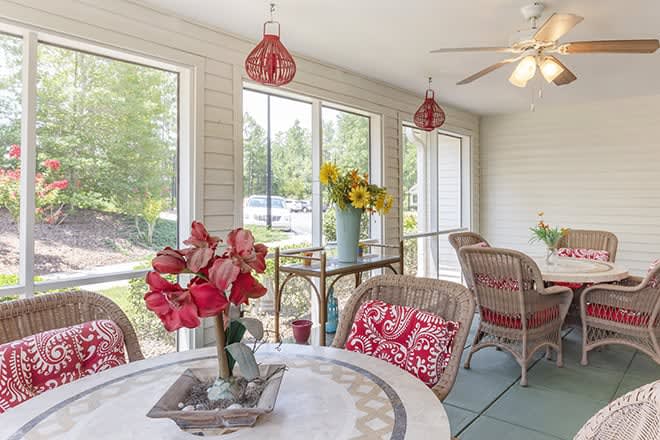 A bright sunroom with wicker furniture, red floral accents, and large windows letting in natural light.