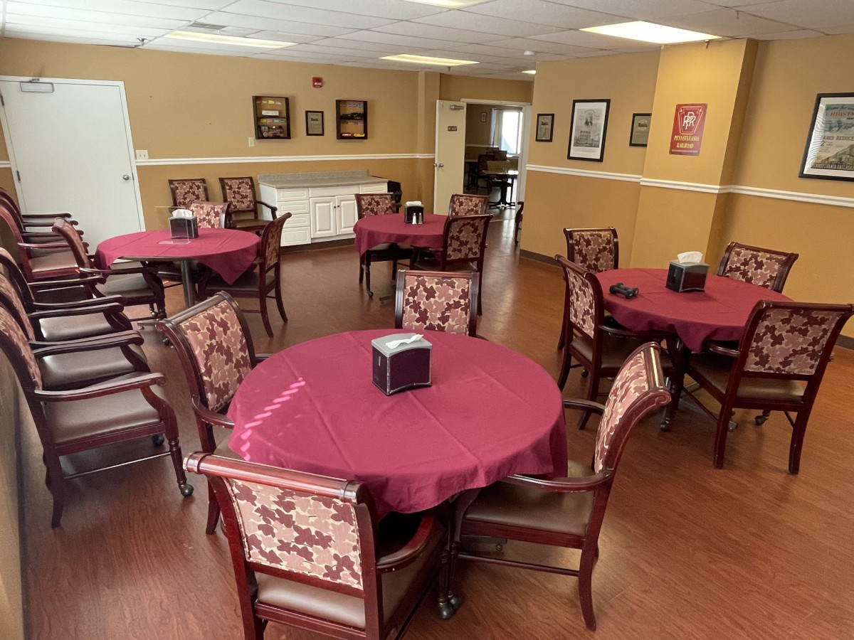 A neatly arranged dining area with round tables and upholstered chairs, each table covered with a maroon tablecloth and a napkin holder at the center.